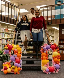 A group of women stand on steps in a library, posing with the number 30 made of balloons