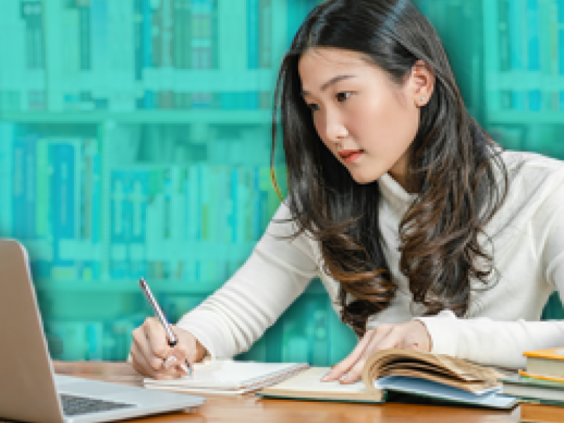image of a woman using a laptop in a library, writing notes
